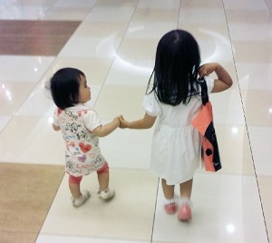 2人の小さな子供が手をつなぎ、右側の女の子が右肩にバックを持っている写真