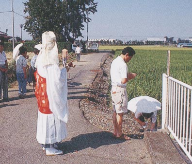 白いフェンスに立てかけられている卒塔婆に向かって手を合わせている男性と僧侶の写真（天明6年水害犠牲者の施餓鬼（木立））