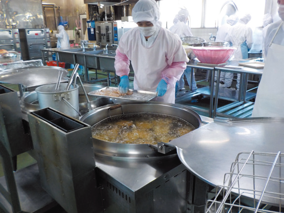 白衣を着た調理員が業務用の大きな鍋を使って給食を作っている写真
